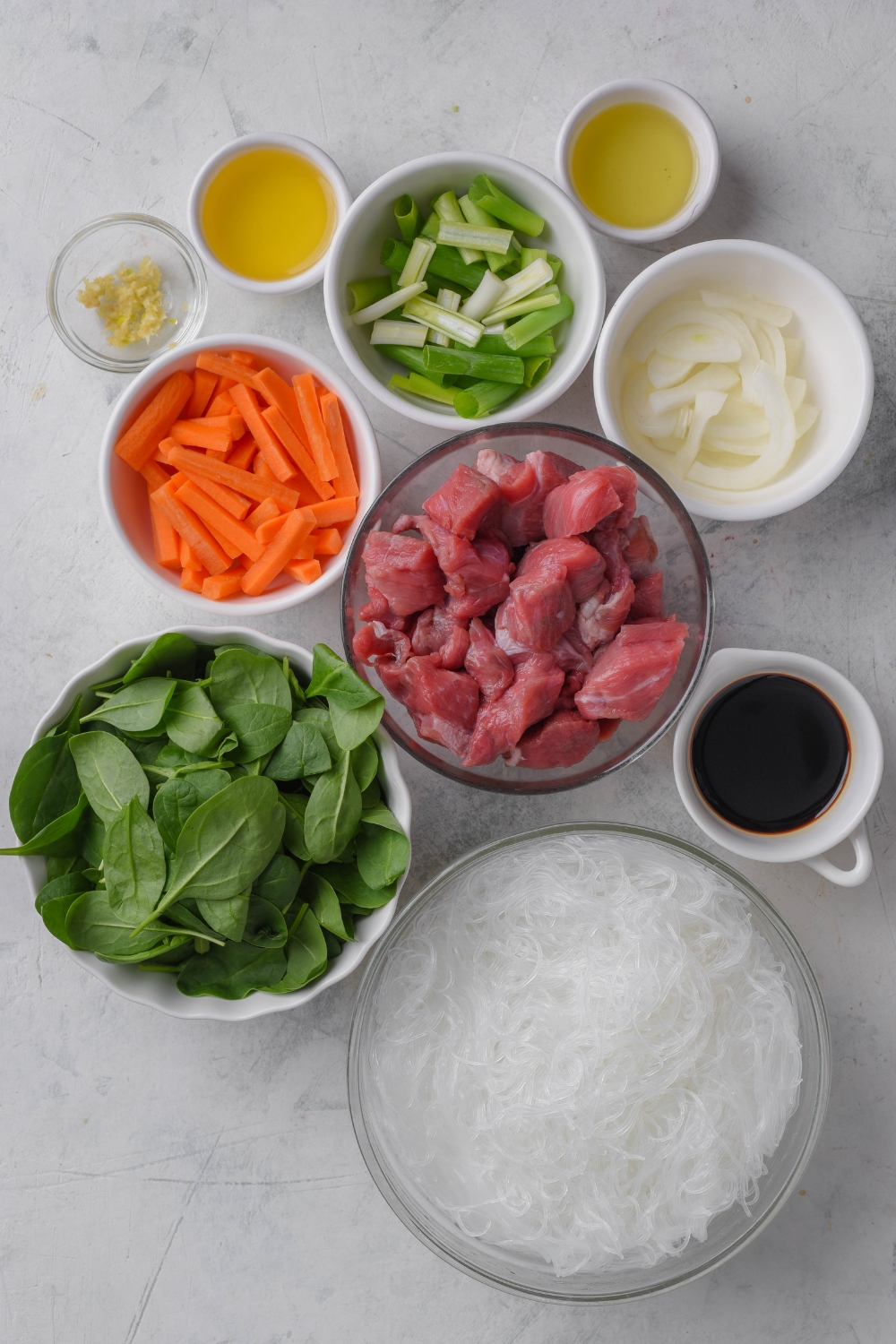A countertop with various bowls containing each ingredient separately. Spinach, beef, glass noodles, soy sauce, carrots, onions, green onions, sesame oil, olive oil, and minced garlic.