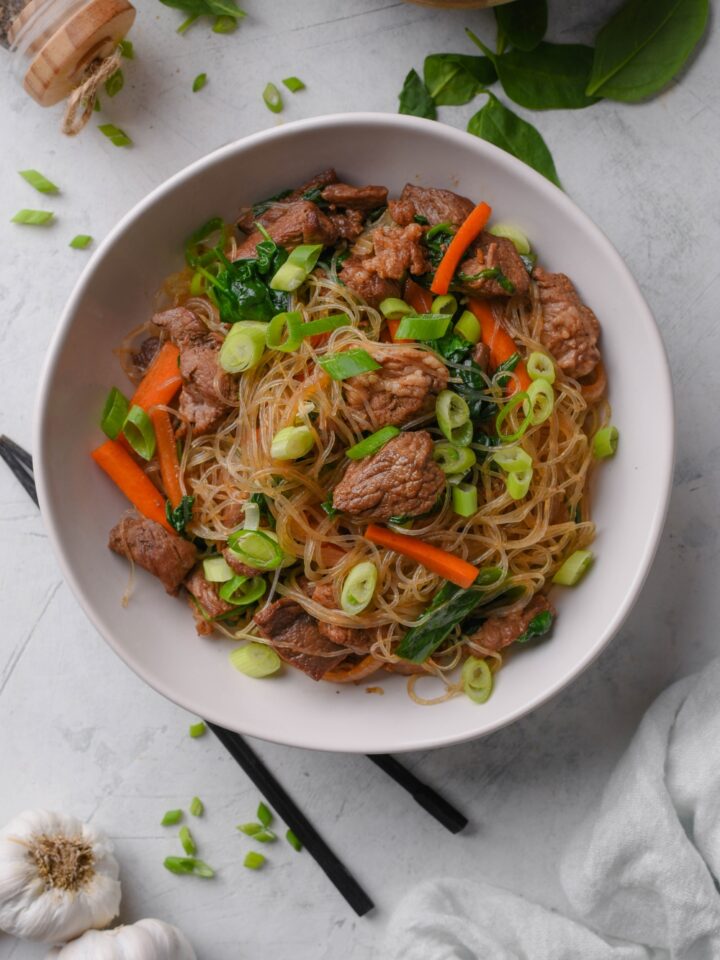 A bowl with glass noodles, beef cubes, green onions, and veggies.
