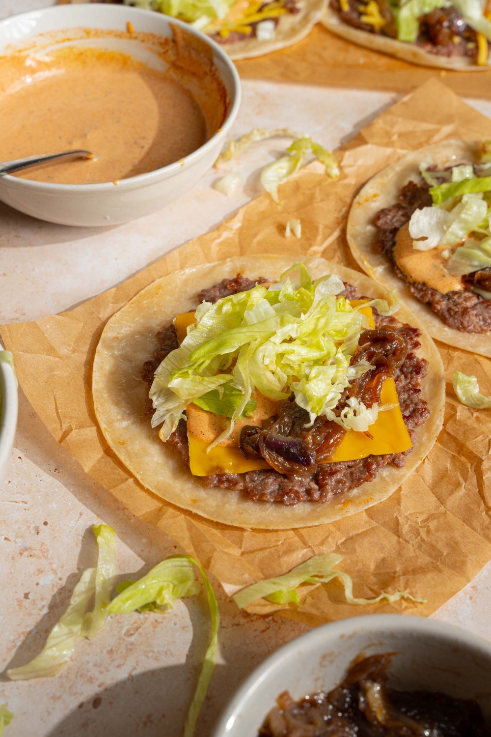 Shredded lettuce on top of smash burger sauce, cheese, and a smash burger all on a flat tortilla on a piece of parchment paper. Next to it is another smash burger taco and behind that is a bowl of smash burger sauce.