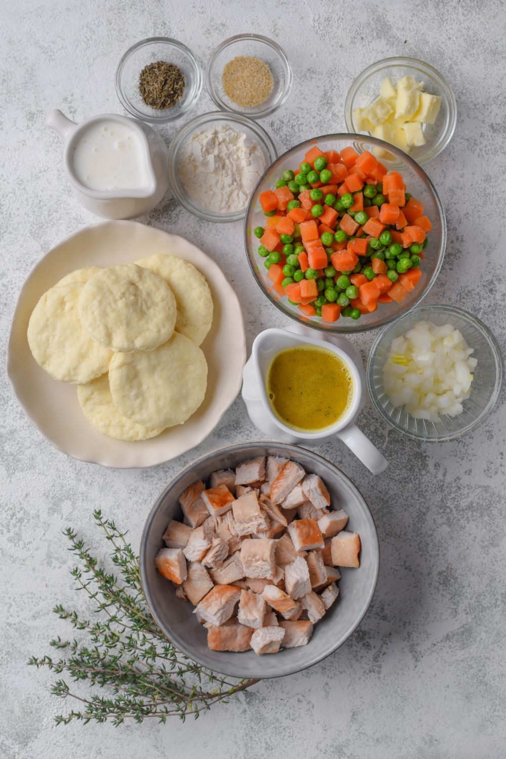 A bowl of chopped turkey, a pitcher of vegetable broth, a bowl of chopped onions, a bowl of biscuits, a bowl of carrots and peas, a bowl of flour, a bowl of butter, a bowl of seasoning, and a pitcher of milk all on a grey counter.