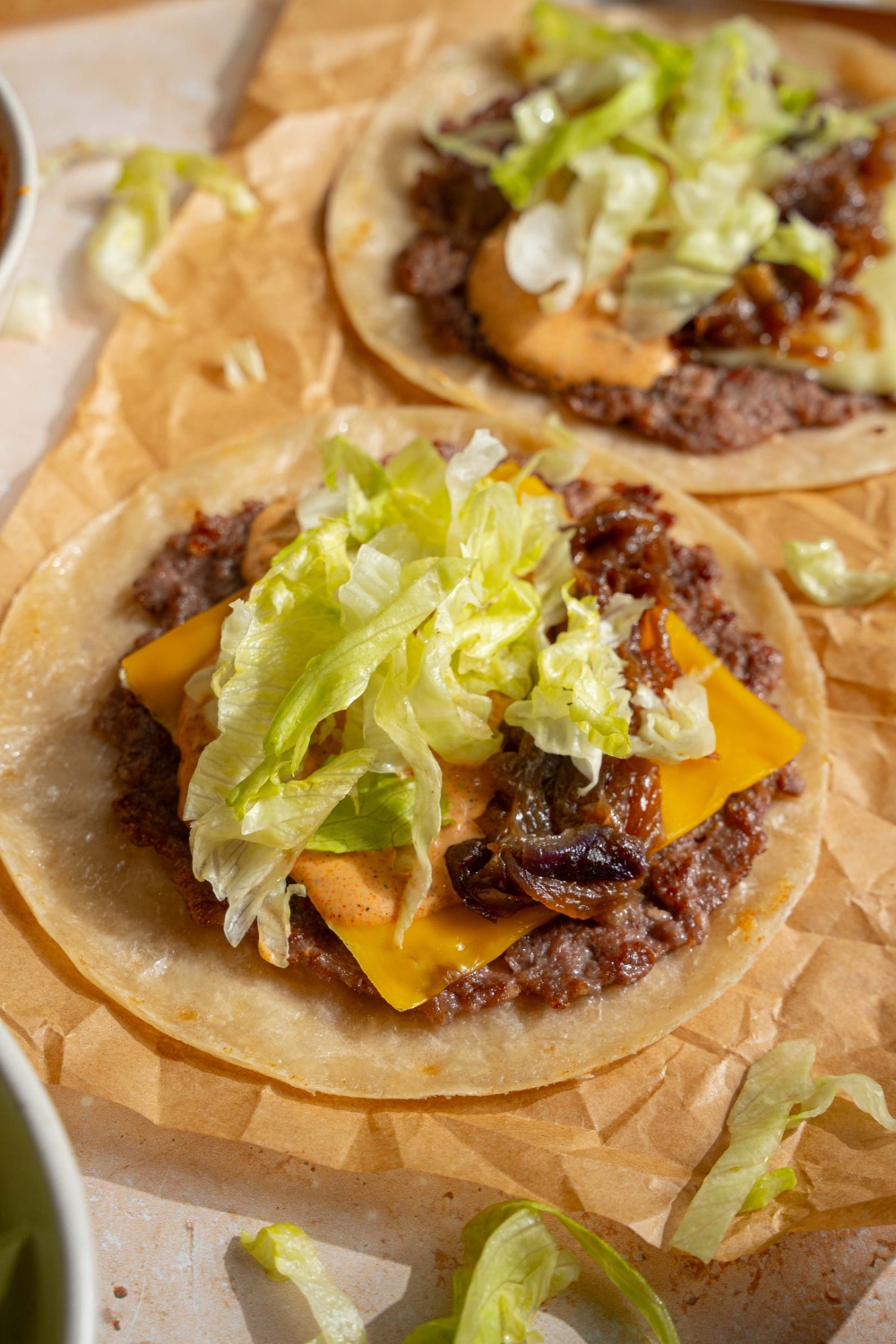 Two smash burger tacos with shredded lettuce, cheddar cheese, smash burger sauce, and a smash burger on a tortilla on a sheet of parchment paper.