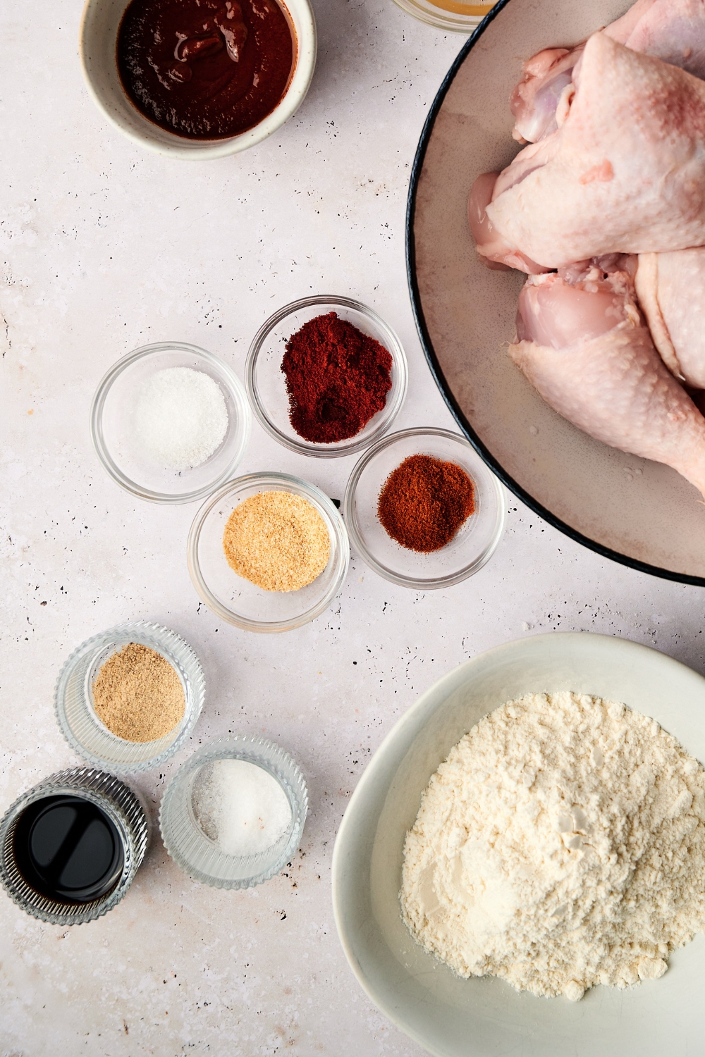 Hot sauce, raw chicken pieces, seasonings, and flour are all in separate bowls on a white counter top.