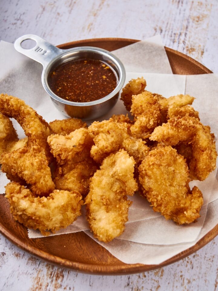 A pile of golden brown shrimp are on a plate lined with paper towels. There is a cup of hot sauce on the plate.