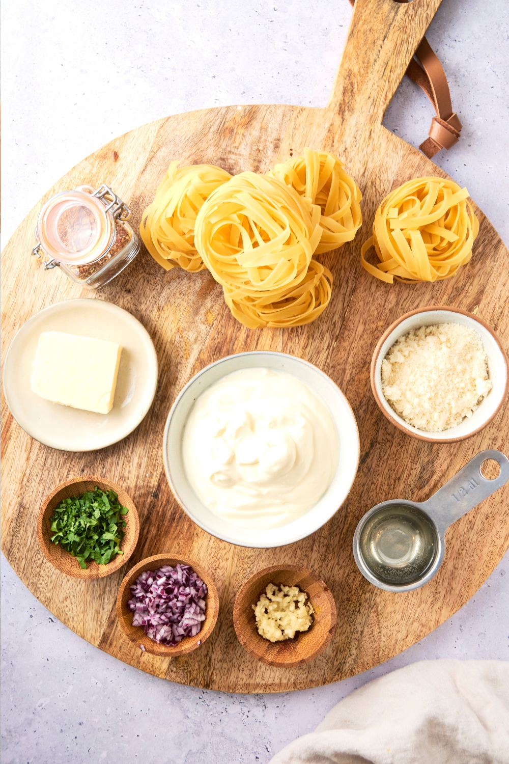 Fettuccine, parmesan cheese, butter, parsley, shallots, garlic, white wine, and oil are all in separate bowls on a wooden cutting board.