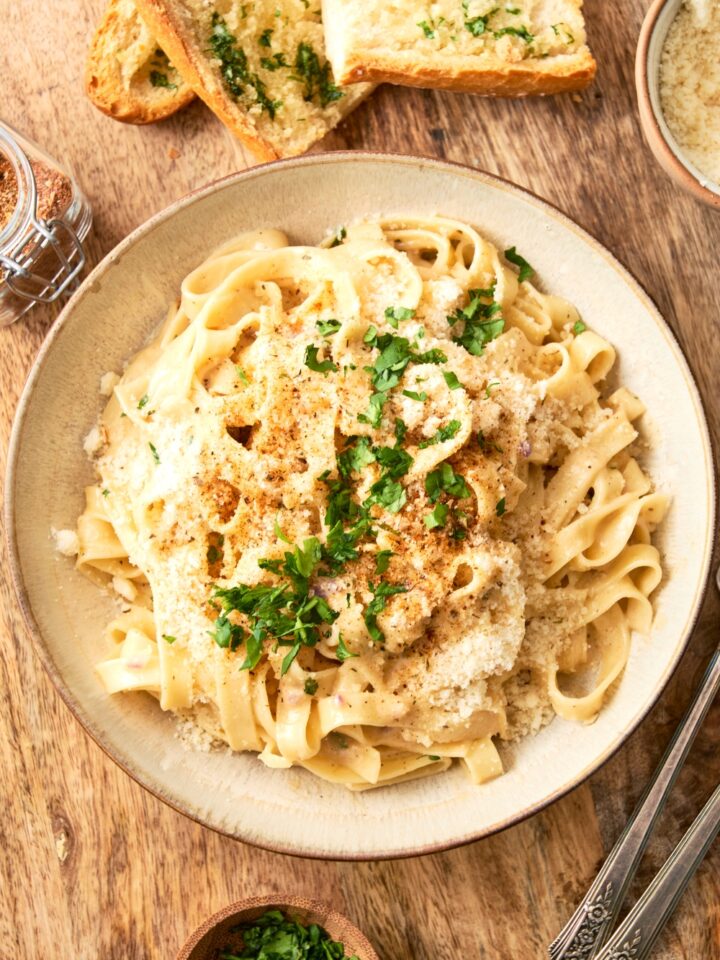 A white bowl is full of fettuccine Alfredo topped with fresh herbs and extra cheese.