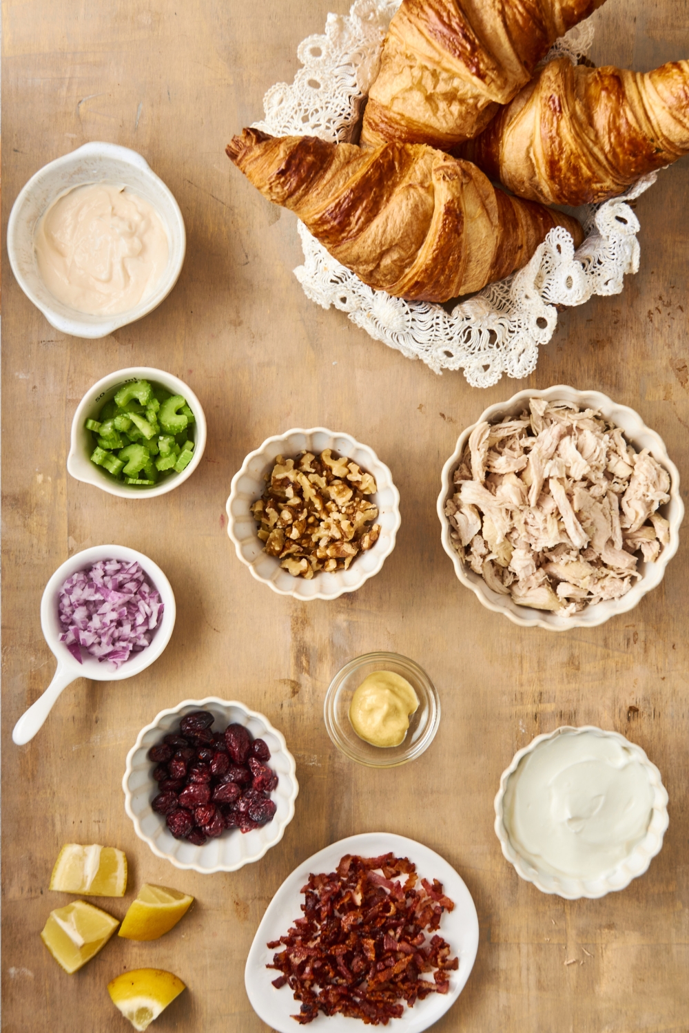 A basket of croissants, and separate bowls holding diced chicken, nuts, celery, red onion, mayo, yogurt, bacon, and cranberries.