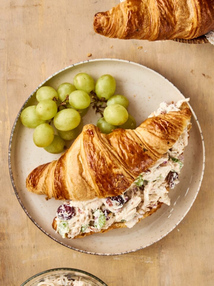 A gray plate holds an overflowing chicken salad croissant and a bunch of green grapes.