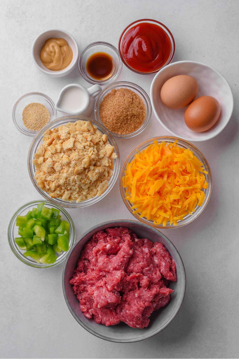 A countertop with shredded cheese, ground beef, diced bell pepper, crushed Ritz crackers, brown sugar, milk, onion, mustard, ketchup, Worcestershire sauce, and eggs in separate bowls.