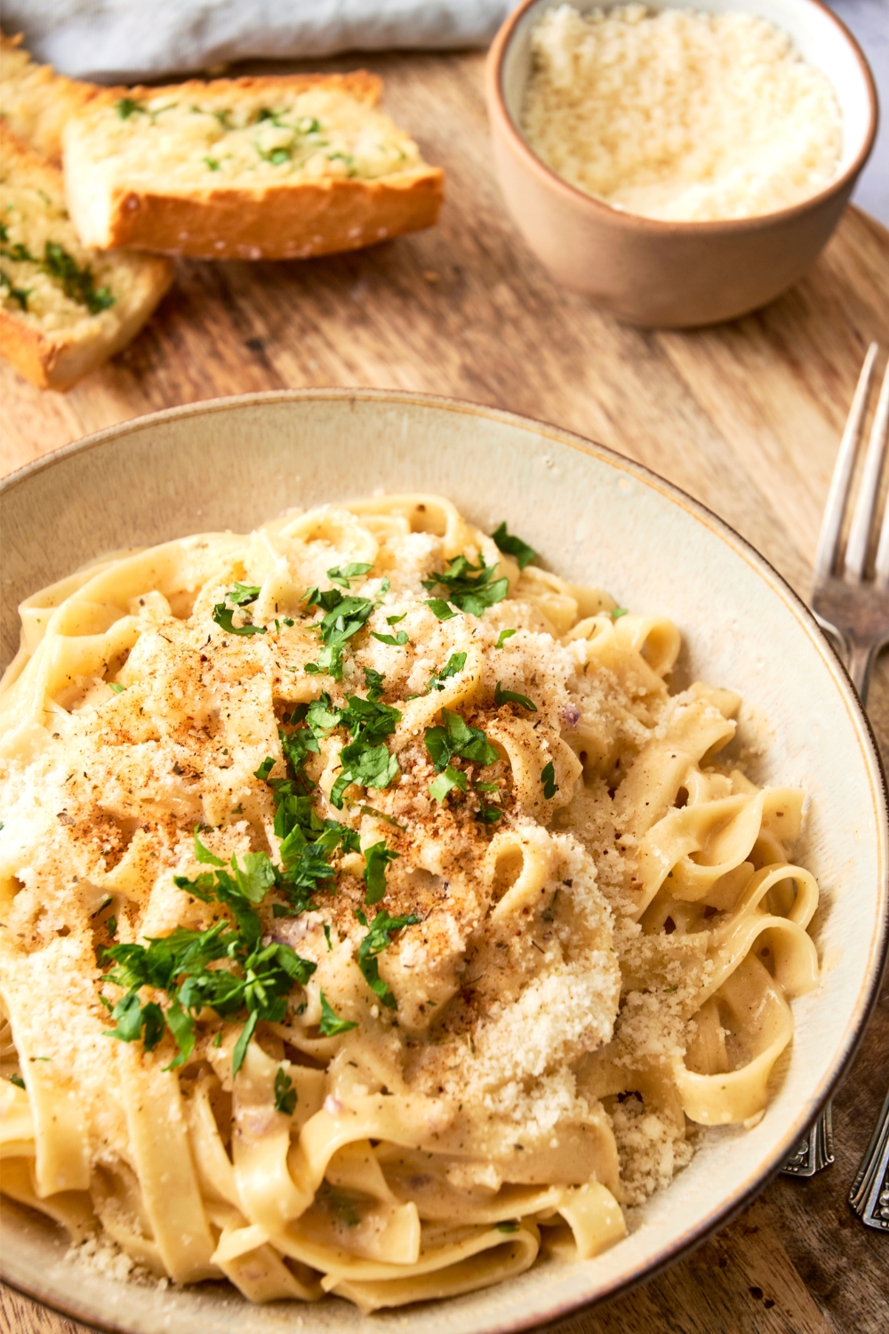 A bowl full of Fettuccine covered in Cajun Alfredo sauce is in a white bowl on a wooden table. A bowl of cheese is in the background and fork rests nearby.