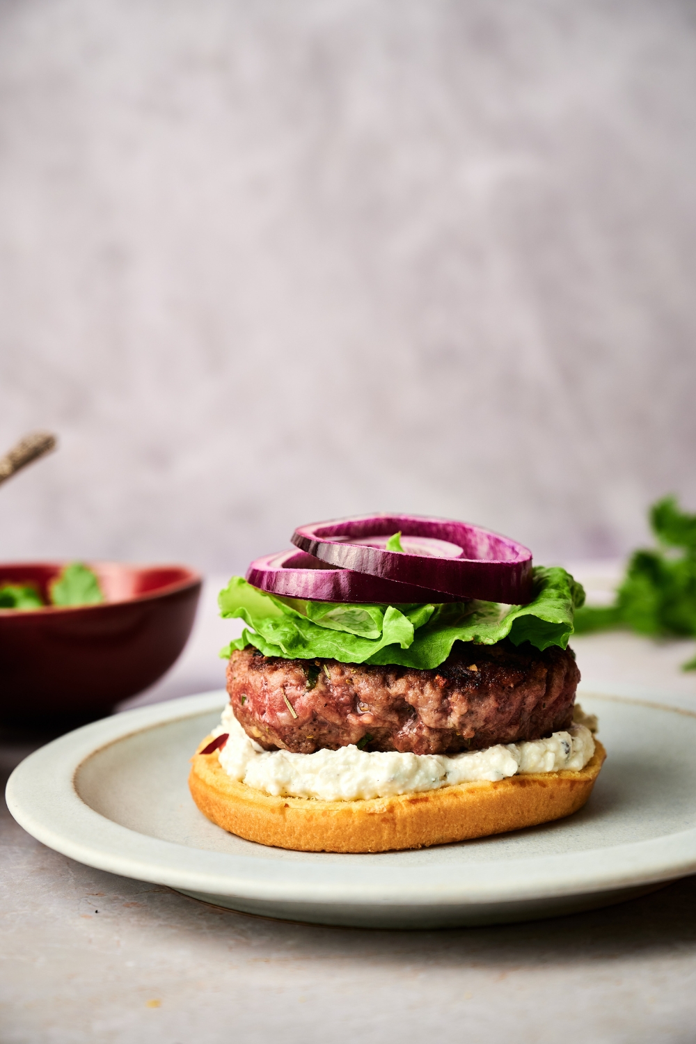 A lamb burger without a top bun sits on a white plate. The burger is topped with onion, lettuce, and feta spread.