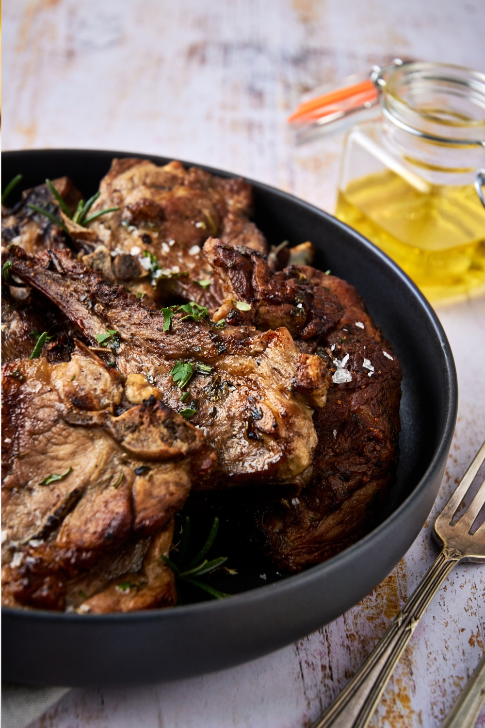 A black dish holds a pile of lamb shoulder chops garnished with fresh herbs and sea salt.