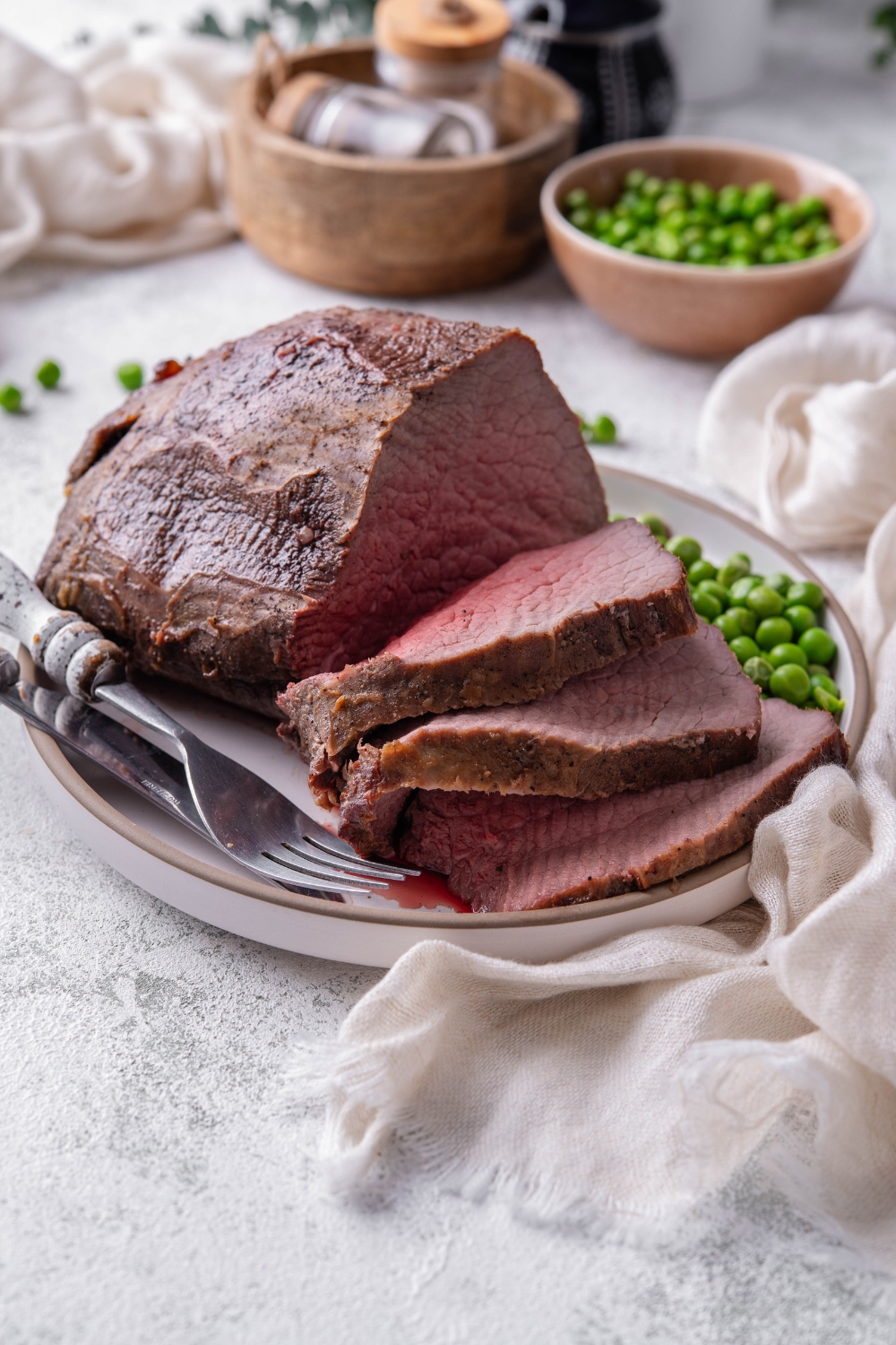 A roast is on a white plate with garden peas and silverware.