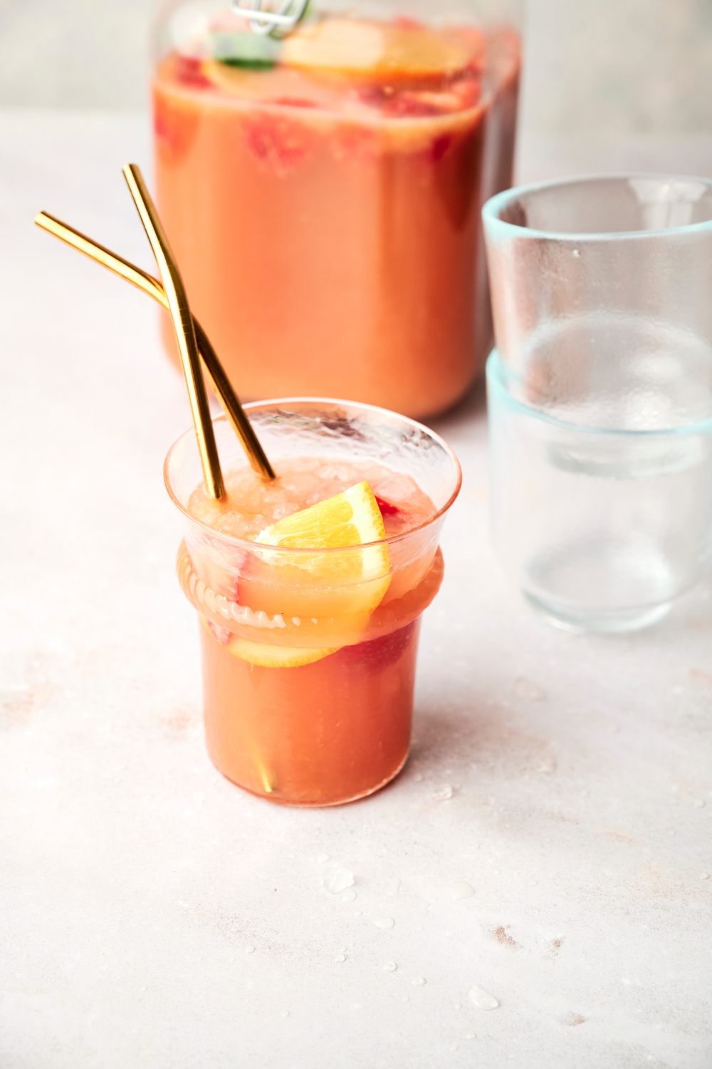 A small glass of jungle juice sits on a counter. It has orange wedges, strawberries, and straws in it.