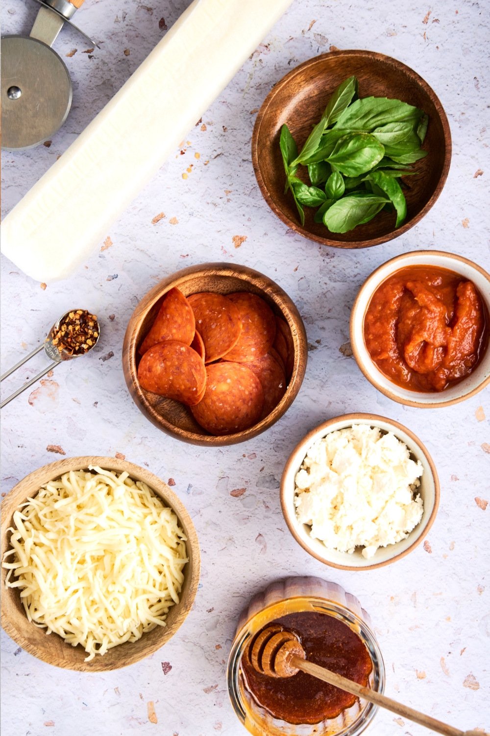 Pizza dough, and bowls with basil, pepperoni, sauce, goat cheese, mozzarella cheese, hot honey, and spices sit on a white counter.