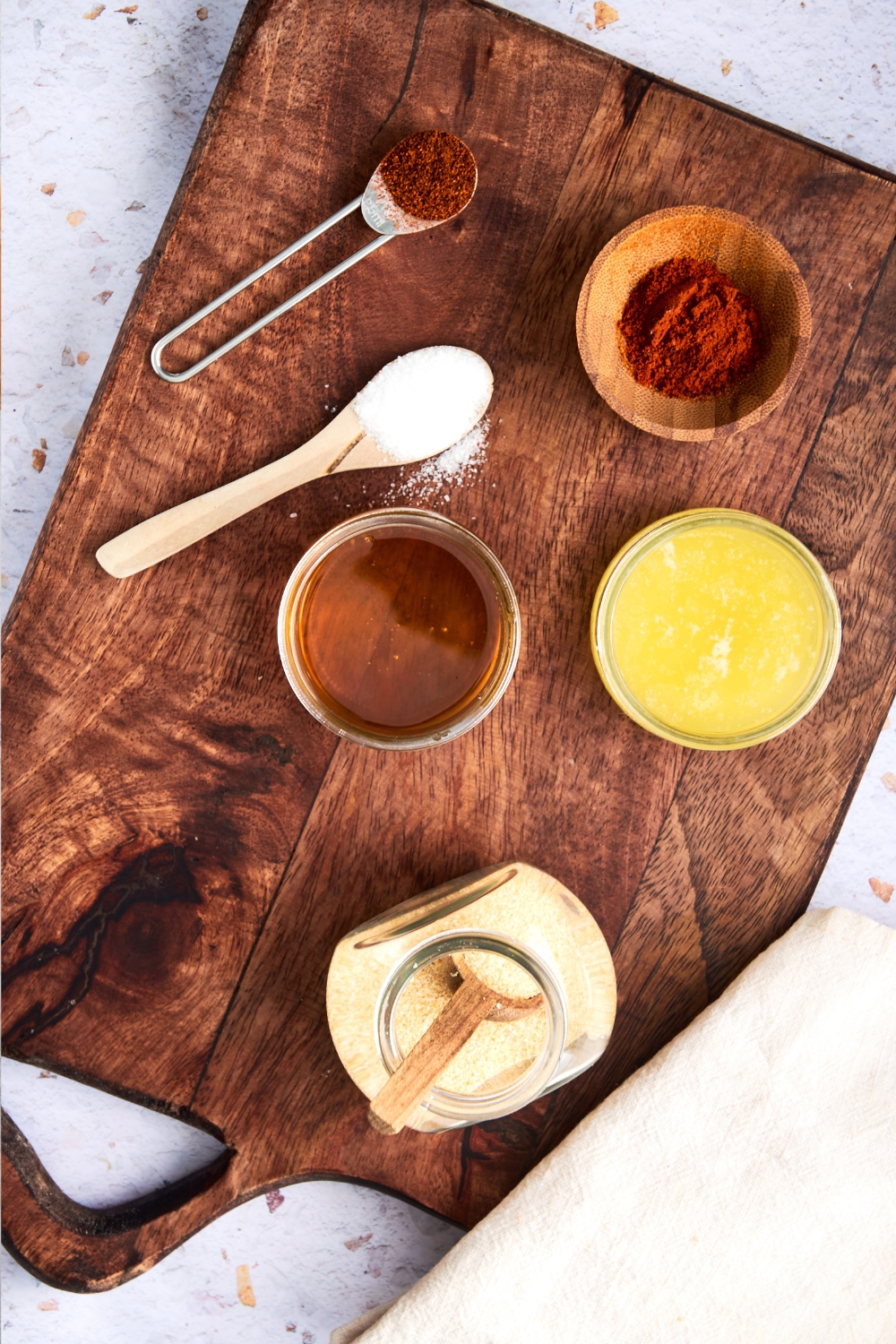 A wooden cutting board holds a glass jar of honey, a jar of melted butter, a wooden bowl of hot paprika, a spoon of chili powder, and a spoon of salt.