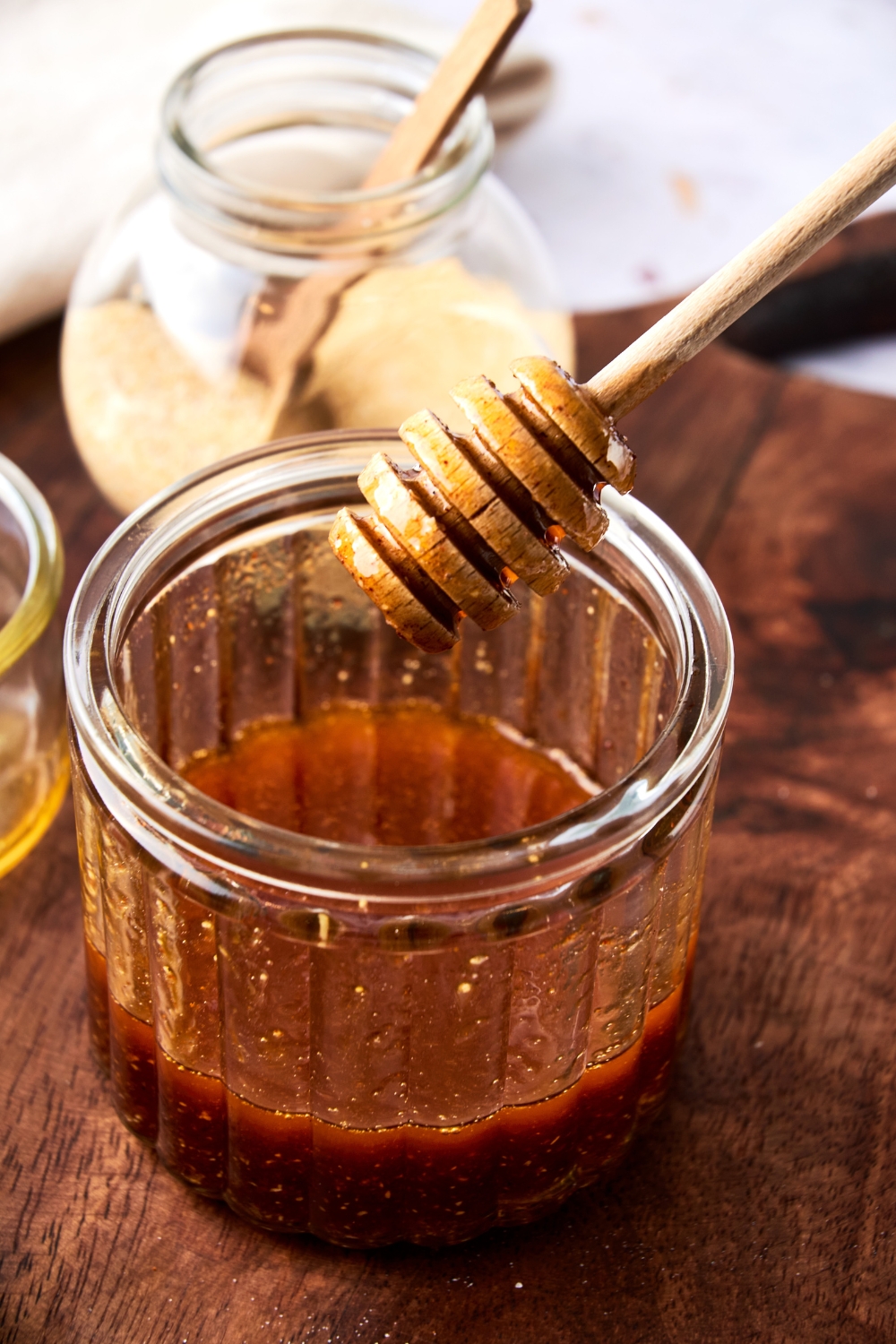 Someone holds a honey dipper of a glass jar full of hot honey on a wooden cutting board.