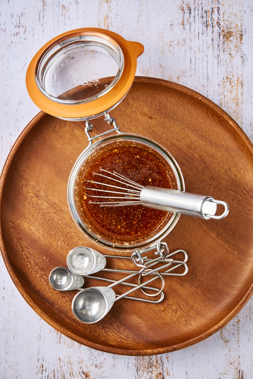 A whisk rests in a glass jar full of Popeye's sweet heat sauce. The jar is sitting on a wooden tray and a set of measuring spoons is near by.