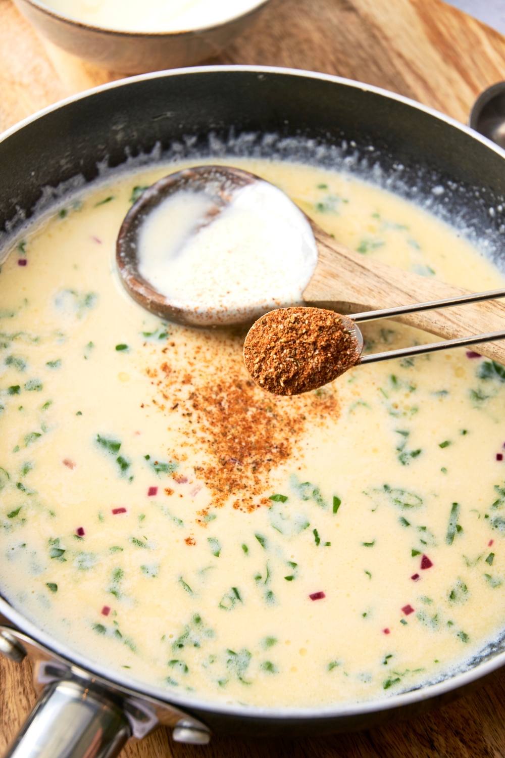 Someone sprinkles Cajun seasoning into a pan full of Alfredo sauce. A wooden spoon rests near by.