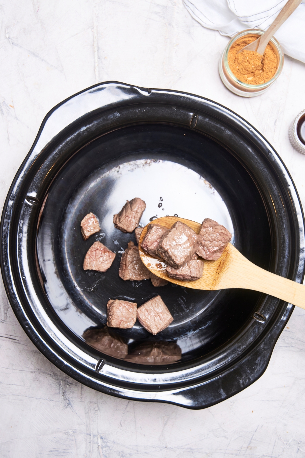 Someone spoons beef into the crock pot using a wooden spoon. A jar of seasoning is near by.