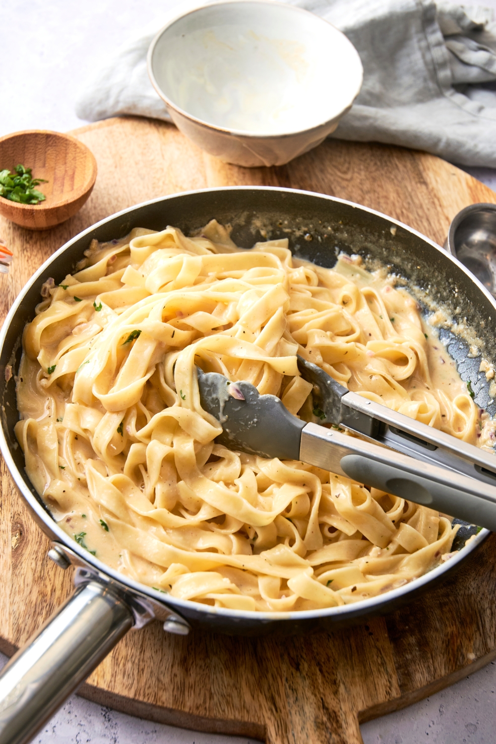 A pair of tongs rest in a pan full of fettuccine covered in Alfredo sauce.