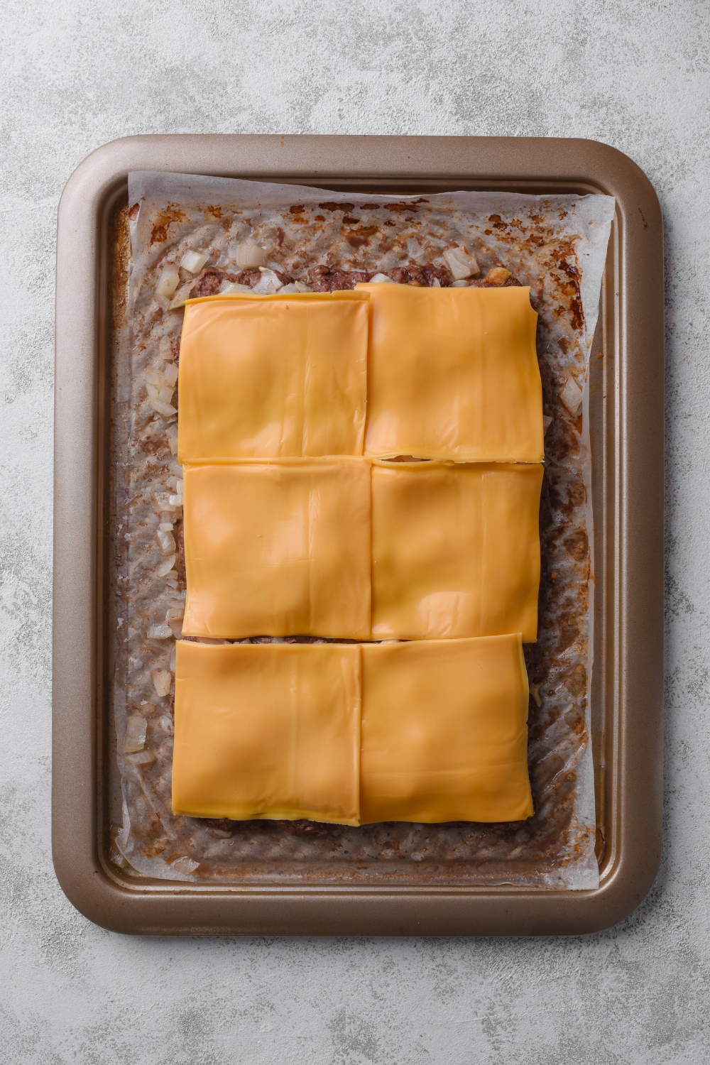 Six slices of cheese cover the ground beef and onions on a parchment paper lined baking tray.