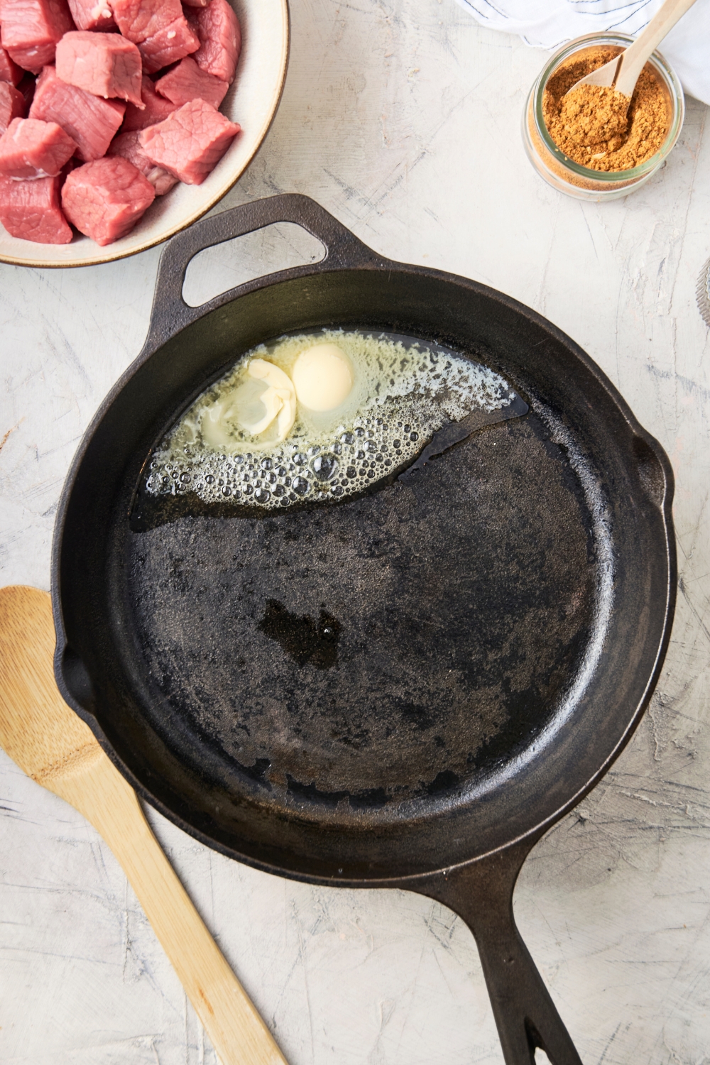 Butter begins to melt in a cast iron skillet. A wooden spoon, seasonings, and raw beef are near by.