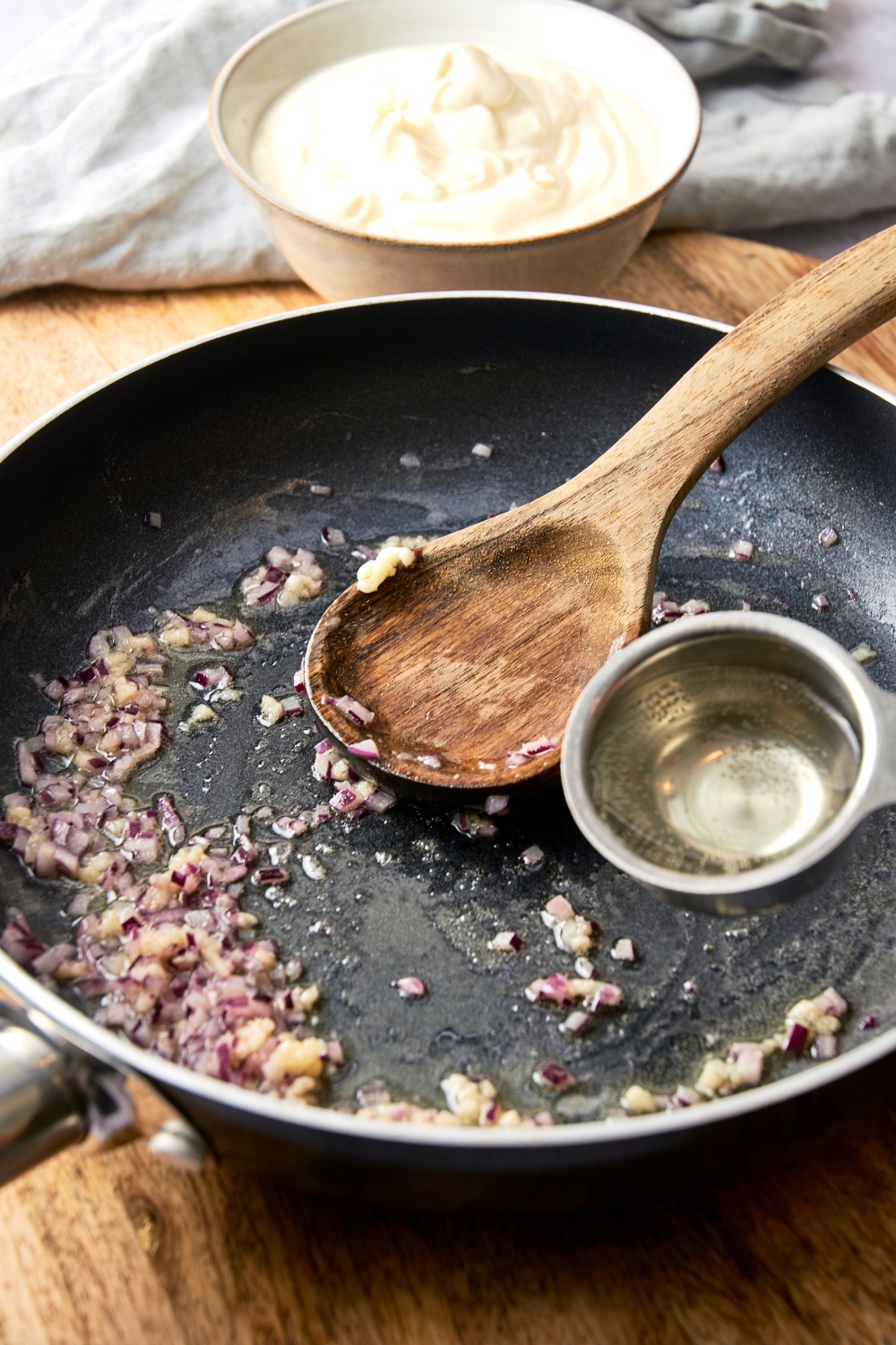 A wooden spoon rests in a black pan with cooked shallots. Someone adds a table spoon of white wine to the pan.