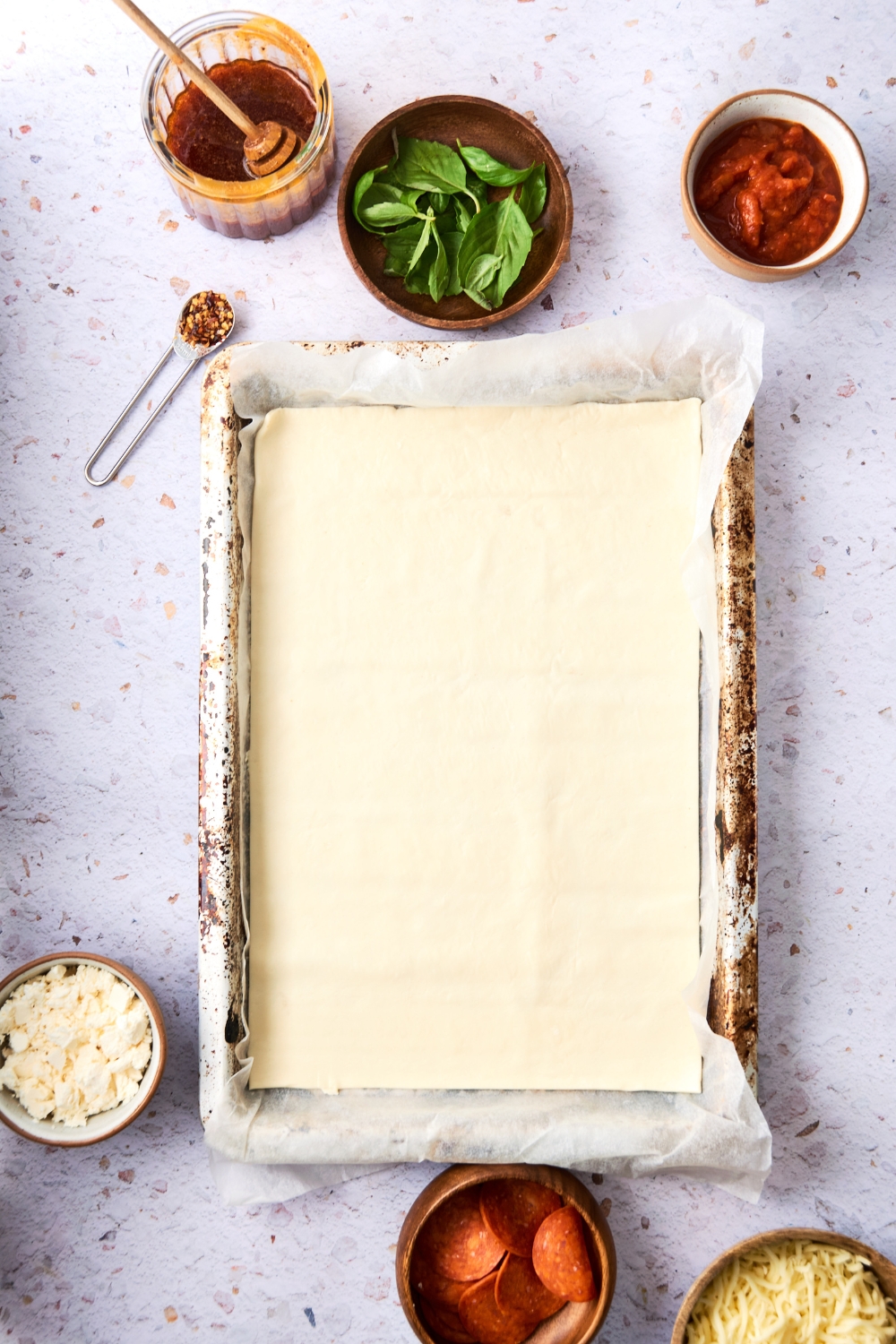 A baking tray lined with parchment paper sits on a counter. There is a pizza crust stretched to fit the tray.