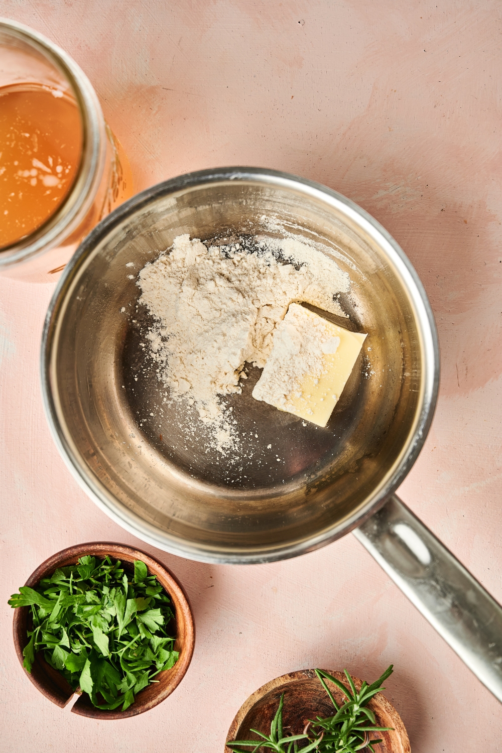 A sauce pan with flour and butter is on a countertop. Wooden bowls of herbs and a glass jar of chicken broth are nearby.