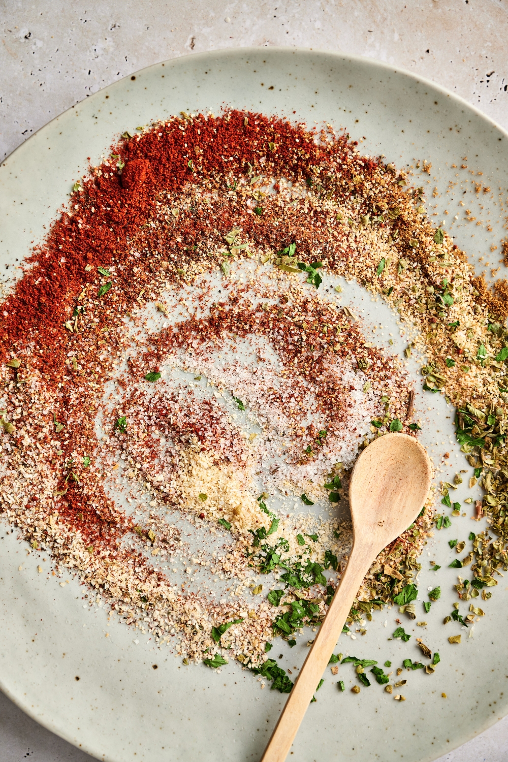 The spices for the cajun two step seasoning have been swirled together on a large gray plate. A wooden spoon is resting in the plate.