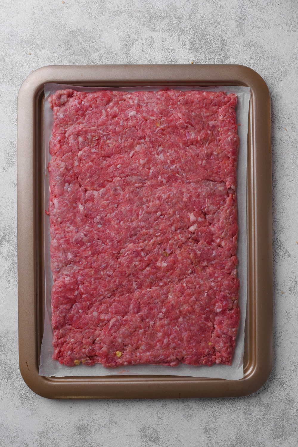 Ground hamburger is spread over a baking tray lined with parchment paper.