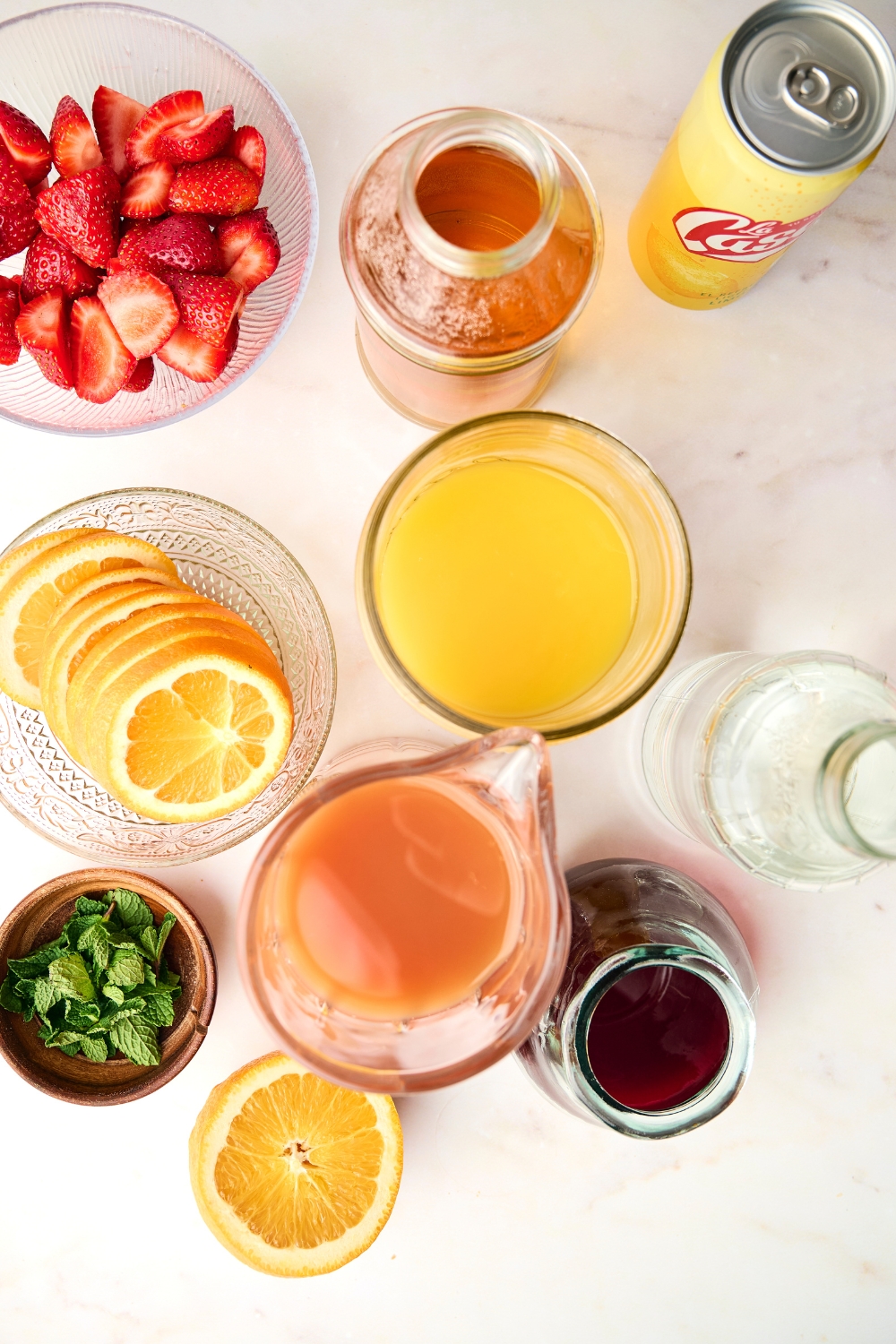 A can of club soda, a bottle of rum, vodka, pitchers of orange and cranberry juices, and a pitcher of fruit punch sit on a counter. Bowls full of strawberries, orange slices, and fresh mint sit near them.