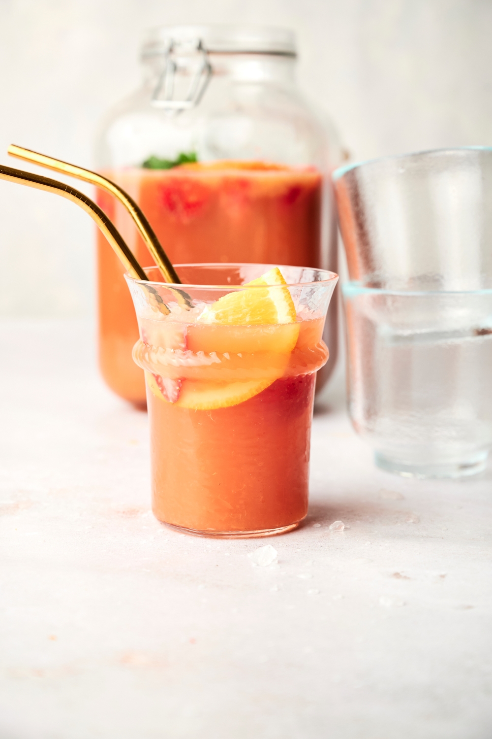 A small glass of jungle juice sits on a counter. There are two golden straws in the glass as well as orange slices.