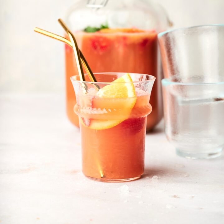 A glass of jungle juice with a slice of orange and strawberries sits on a counter. Two gold straws are sticking out of the cups.