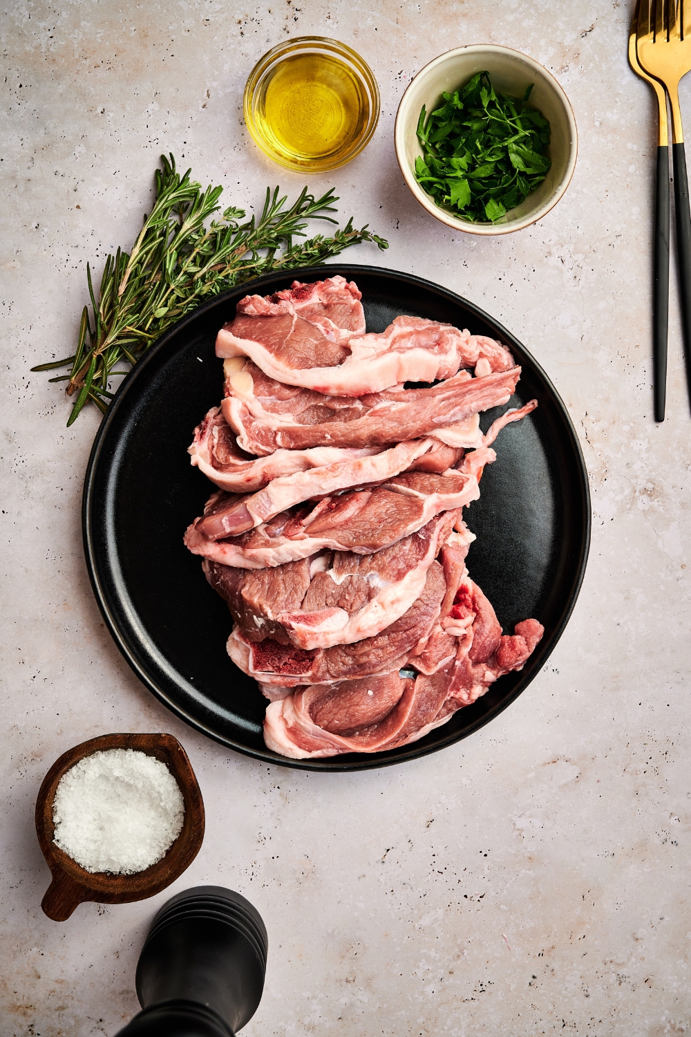 Oil, parsley, rosemary, salt, and lamb lollipops are all in separate containers on a counter top.