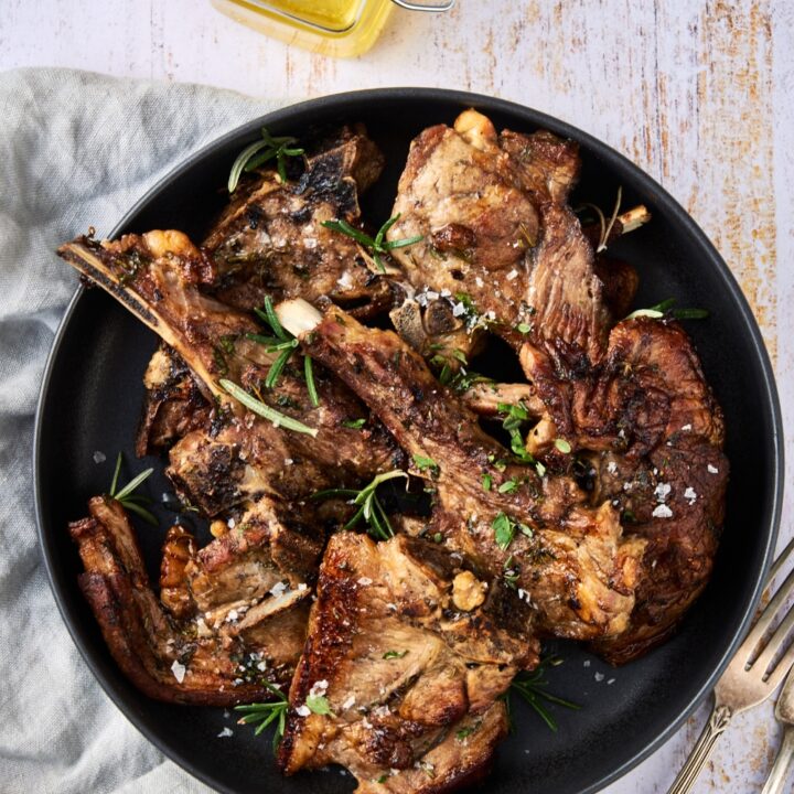 A black dish is full of lamb shoulder chops. The chops are garnished with fresh herbs and sea salt. A fork rests on the counter next to the dish and a jar of oil is nearby.