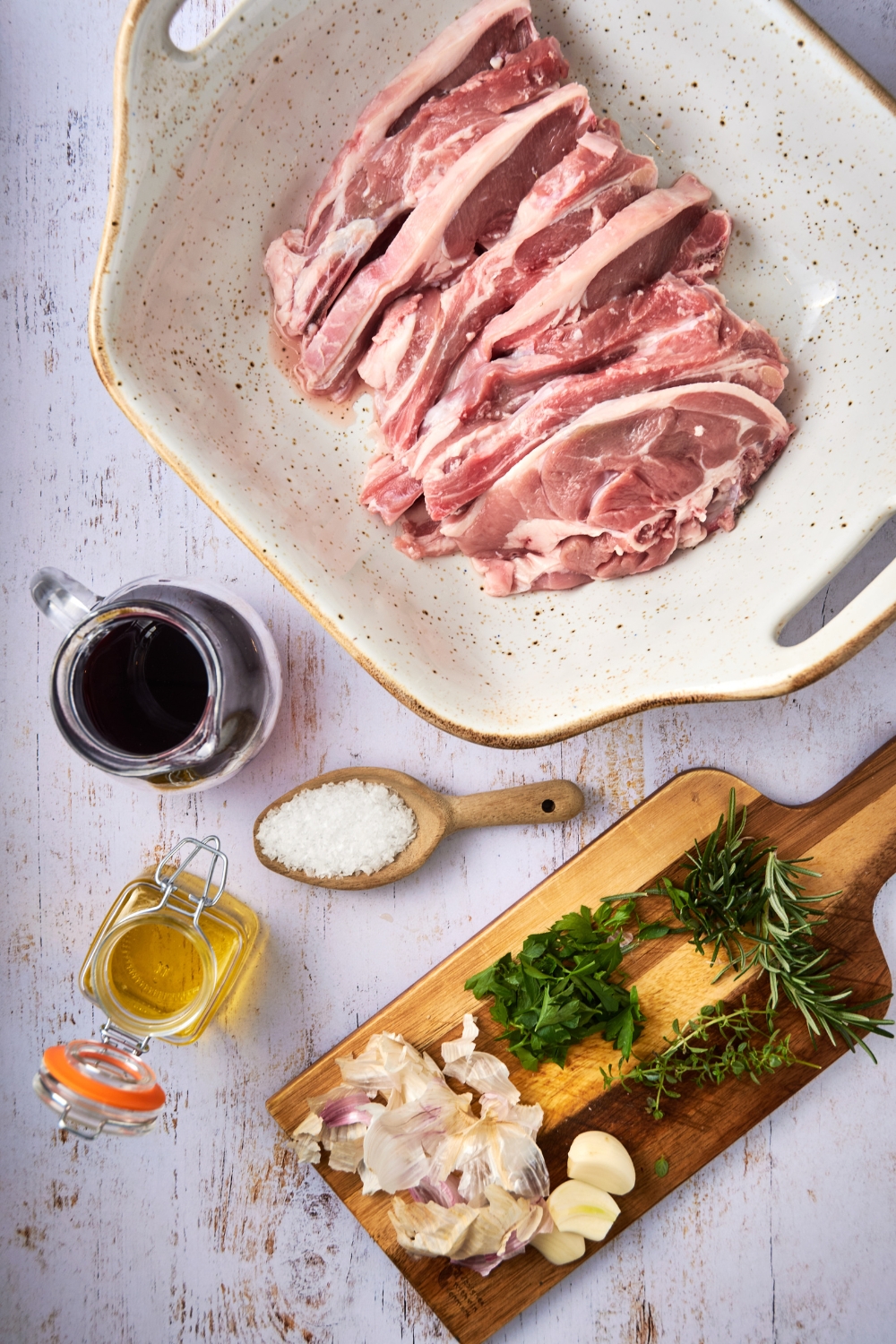 A wooden cutting board holds fresh herbs, sliced shallots, and garlic cloves. A white dish holds raw lamb shoulder chops and oils and salts are in separate containers on a white countertop.