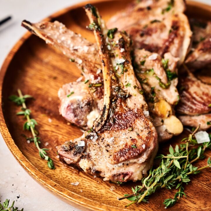 A wooden serving board holds a pile of seared lamb lollipops and fresh rosemary.