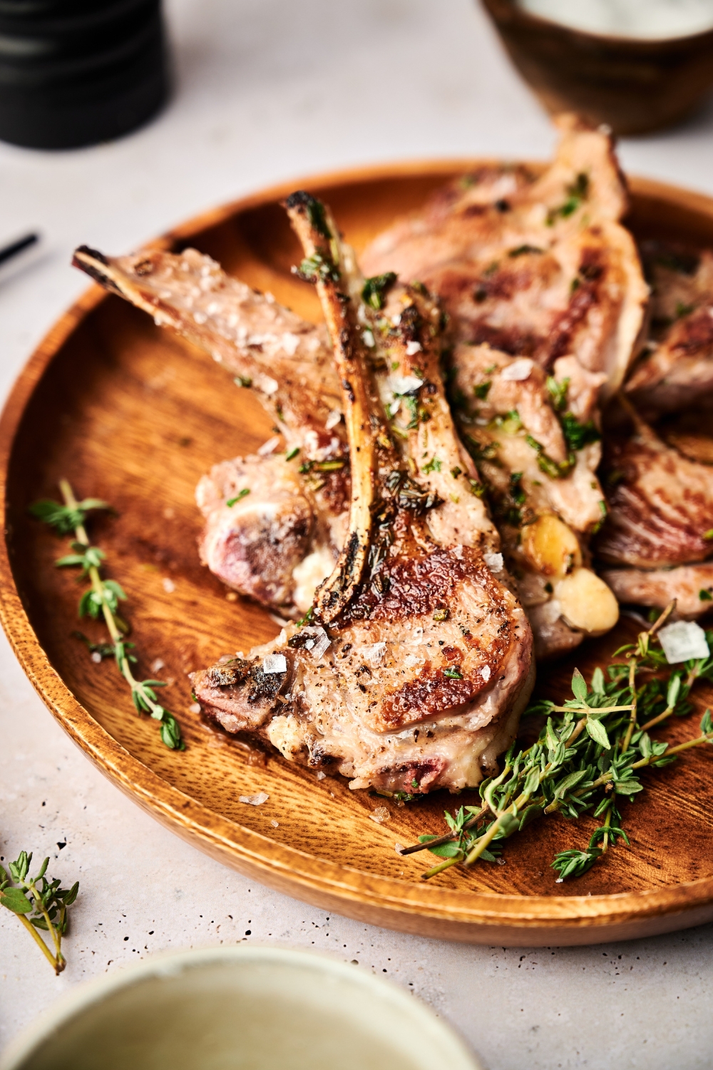 A wooden serving board holds a pile of seared lamb lollipops and fresh rosemary.