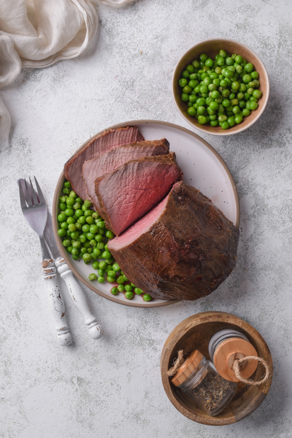 A rump roast sits on a white plate. Silverware is on the counter next to the plate and garden peas are in a bowl next to it.