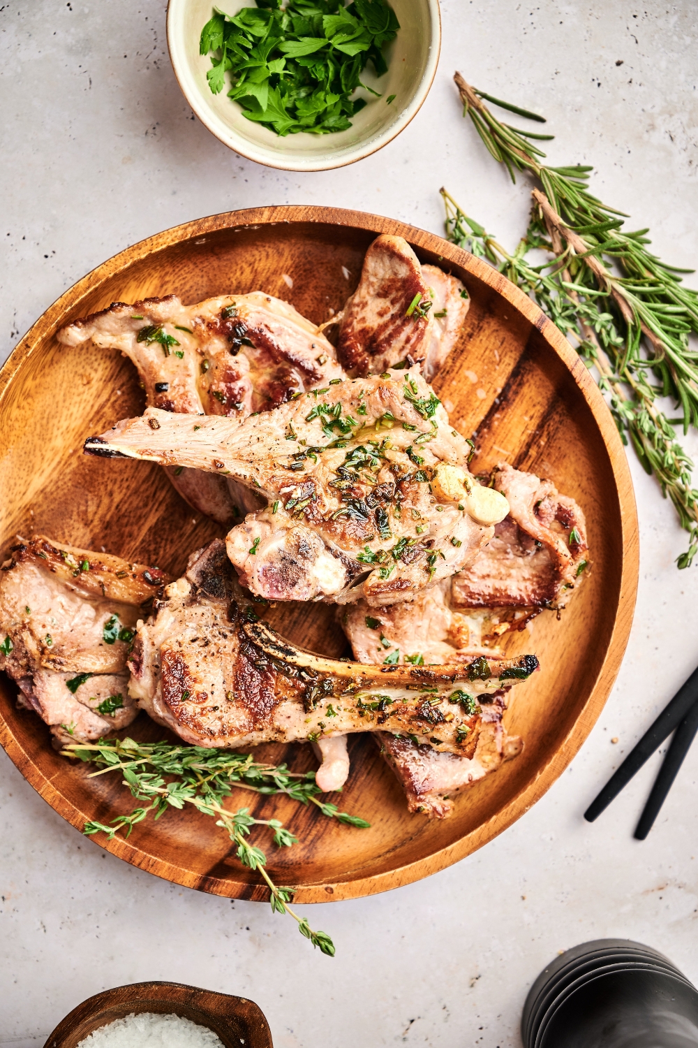 A wooden serving board holds seared lamb lollipops, fresh rosemary sprigs, and sea salt.