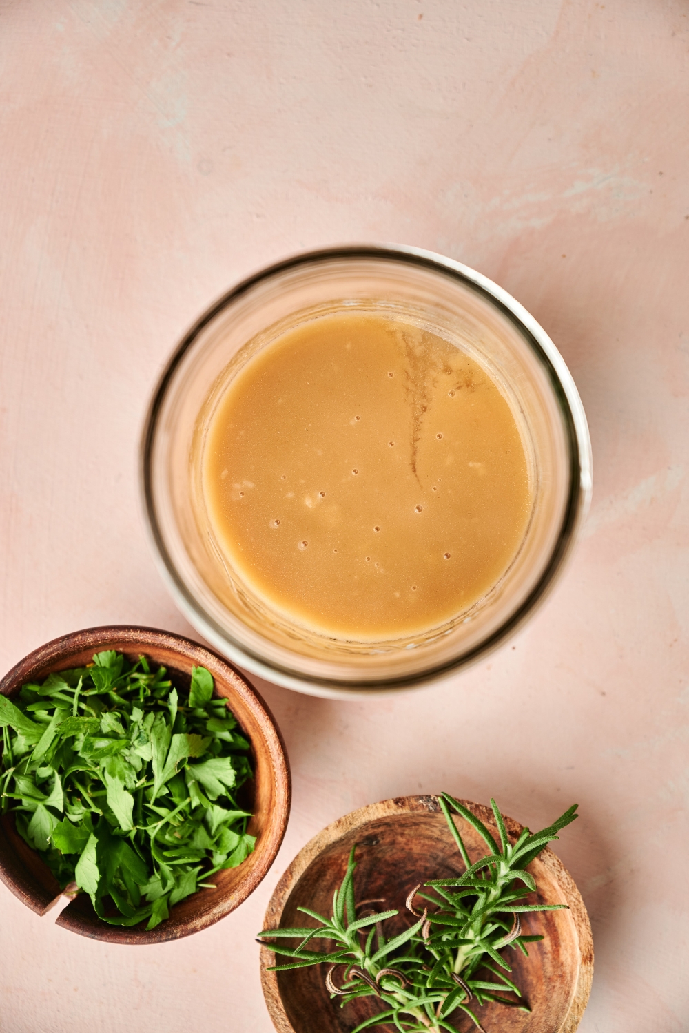 A glass jar full of gravy sits on a counter next to wooden bowls of fresh herbs.