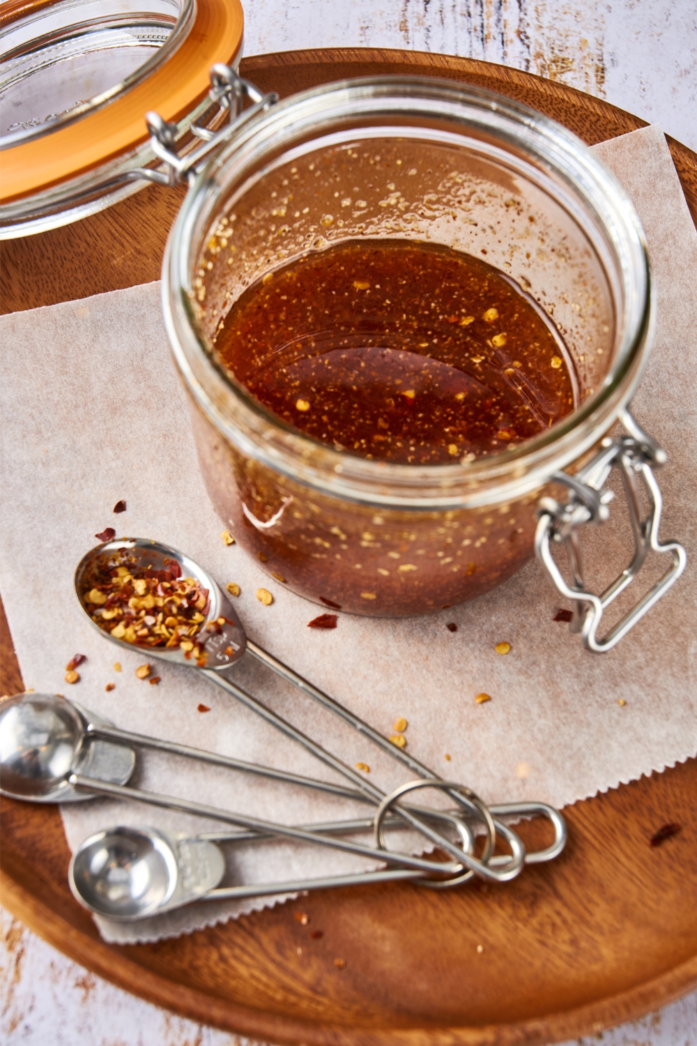 A glass jar full of Popeye's sweet heat sauce sits on a tray. A set of measuring spoons and red pepper flakes are on the tray as well.