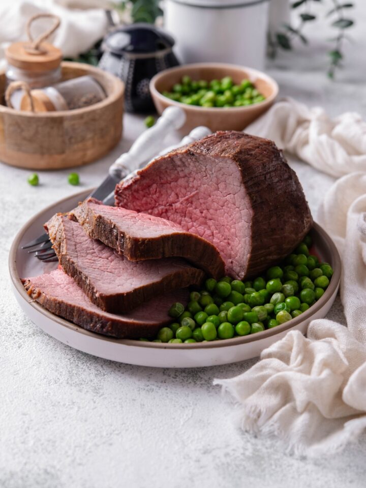A medium rare rump roast sits in a shallow white dish. Garden peas and silverware are on the dish as well.