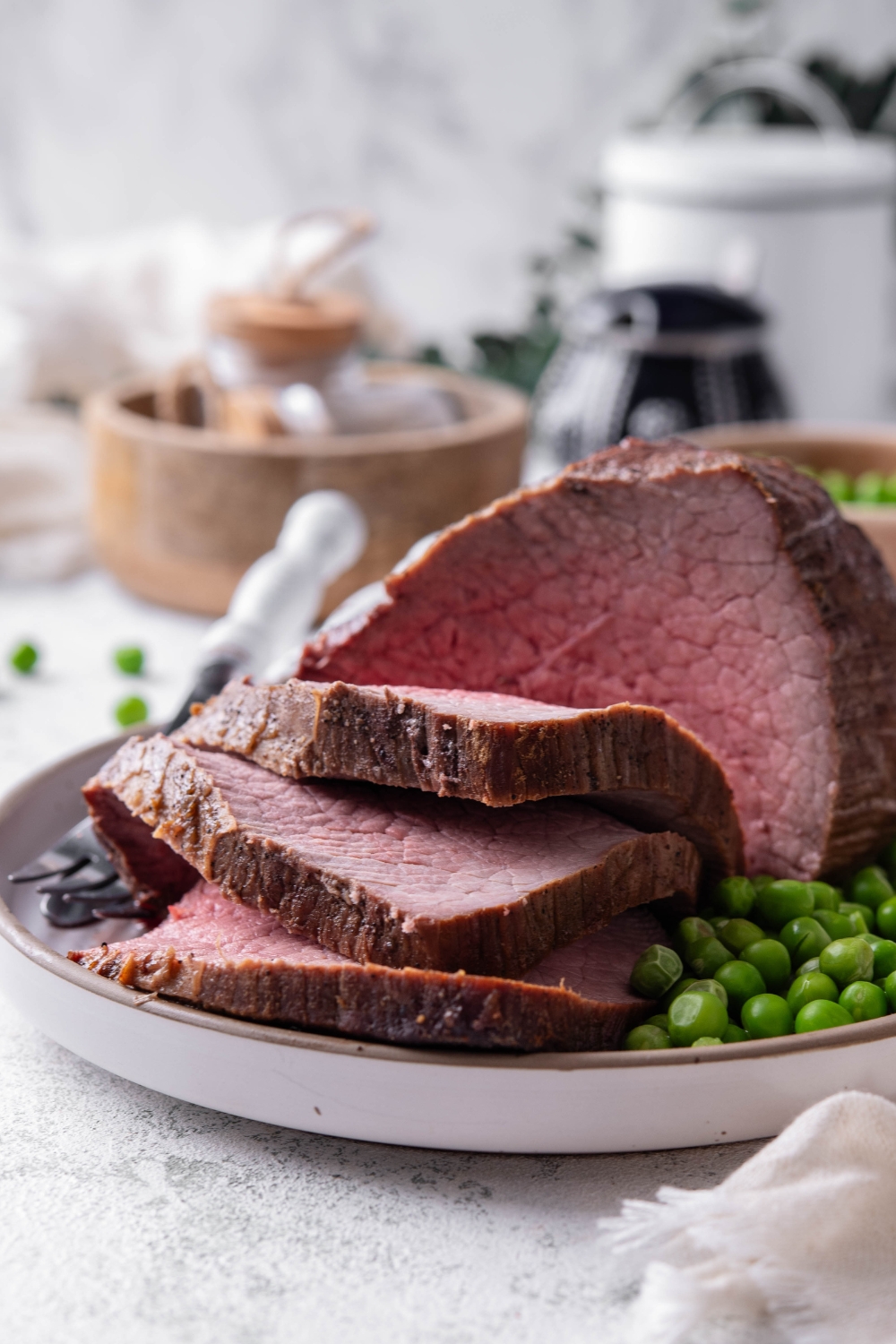 A medium rare rump roast is in a white shallow dish. Three thick slices of roast have been cut from it. Peas and silverware are on the plate.