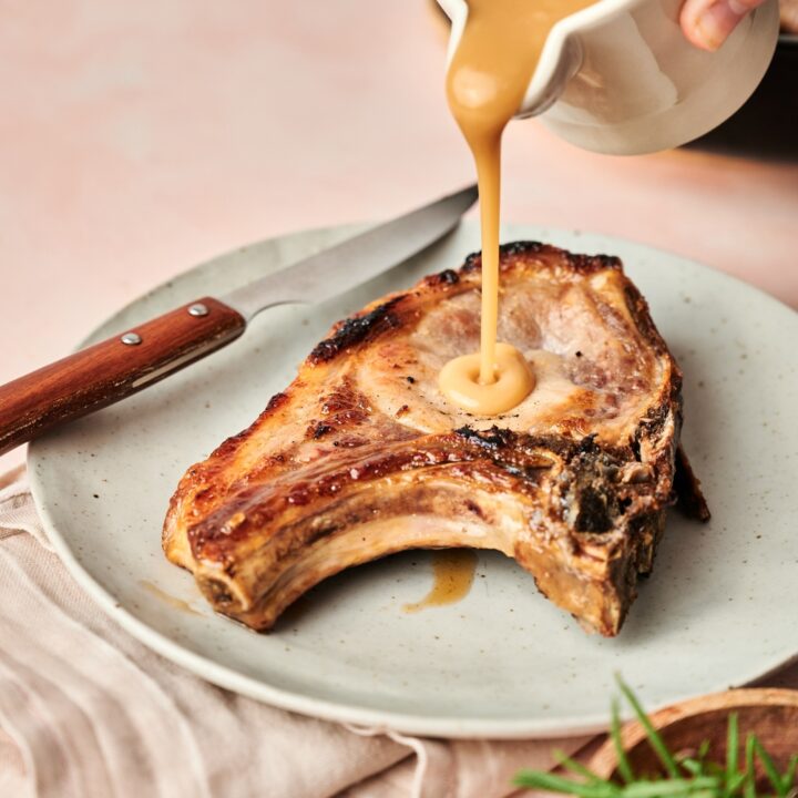 Someone pours gravy over a pork chop on a plate. A knife is resting near by.