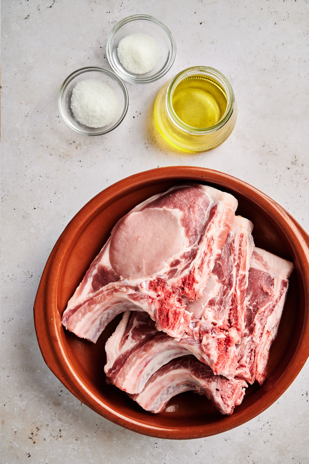 Pork chops sit in a large red bowl and smaller jars of salt, sugar, and oil are next to it.