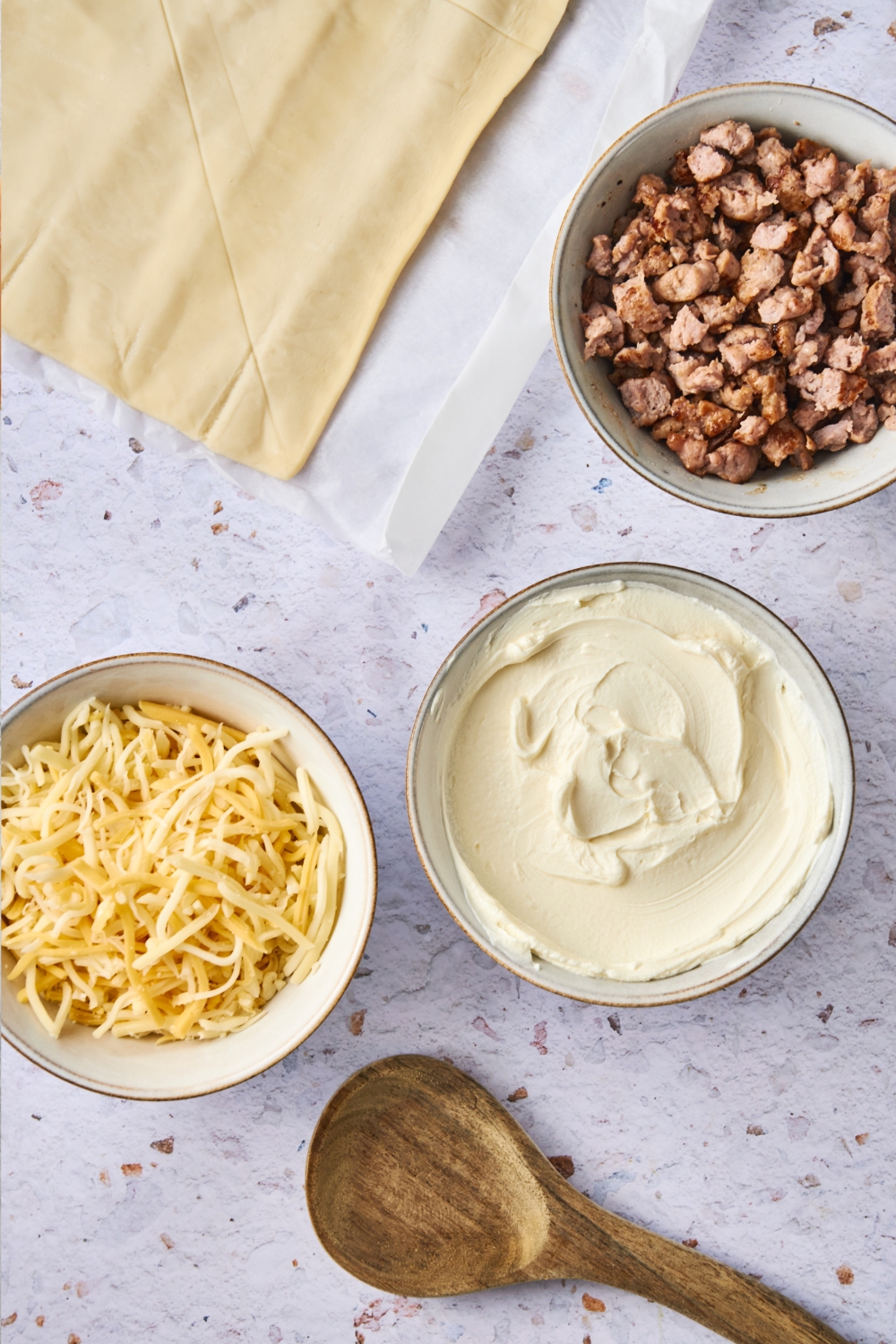 Unrolled crescent dough is on a counter. Bowls with crumbled sausage, cream cheese, shredded cheddar cheese, and a wooden spoon are there as well.