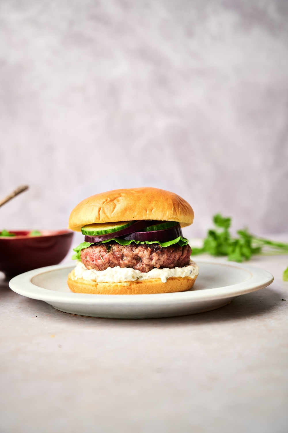 A lamb burger sits on a white plate. The burger is topped with feta spread, lettuce, onion, and cucumber.