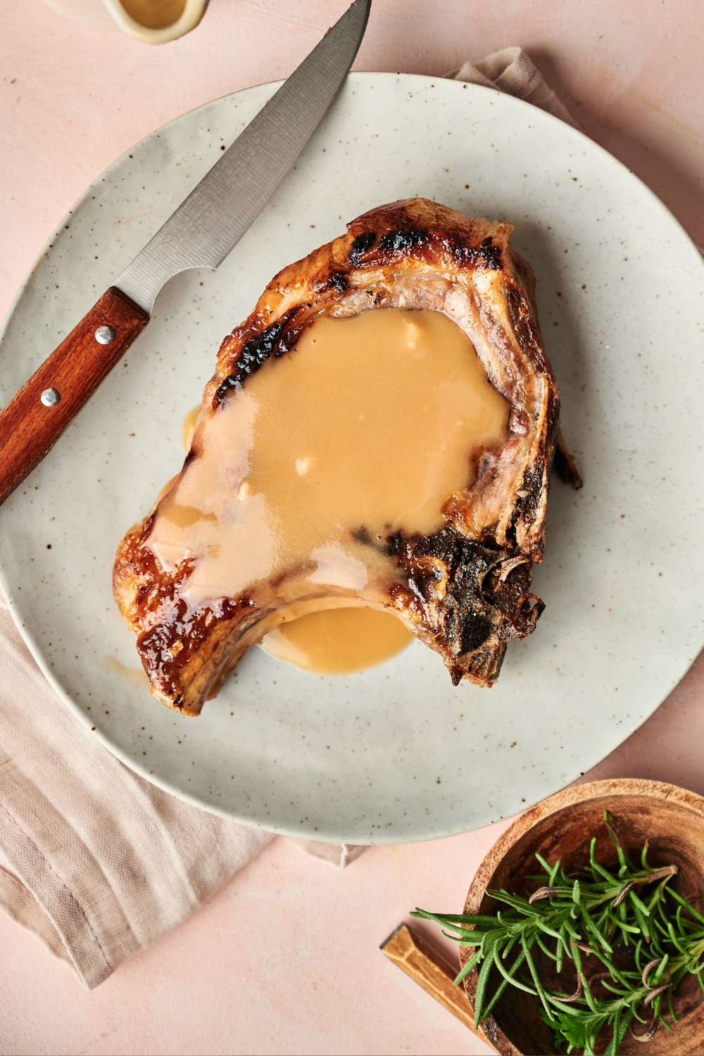 A pork chop sits on a gray plate. The pork chop has been covered in gravy and a knife rests near by.